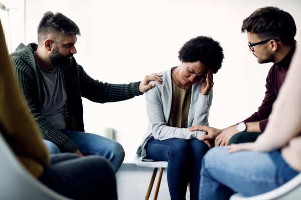 Supportive members of group therapy consoling sad African American woman during psychotherapy.